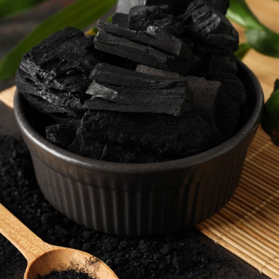 Ciotola nera contenente pezzi di carbone vegetale naturale con polvere di carbone e cucchiaio in legno su superficie in bambù.