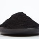 Primo piano di carbone vegetale in polvere fine di colore nero disposto su un piatto scuro con sfondo chiaro.